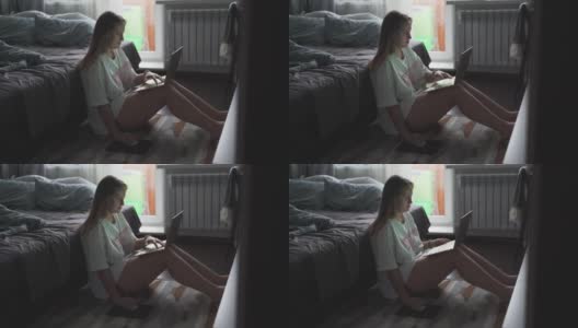 在一个多云的清晨，一名年轻女子正坐在电视机前的地毯上用笔记本电脑工作。她仰靠在摊开的沙发上。在家工作流行病商务距离高清在线视频素材下载