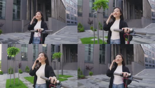 好看的满意的幸运的年轻亚洲女孩在休闲服装享受她的电话交谈，而走在现代建筑附近与自己的电动滑板车高清在线视频素材下载