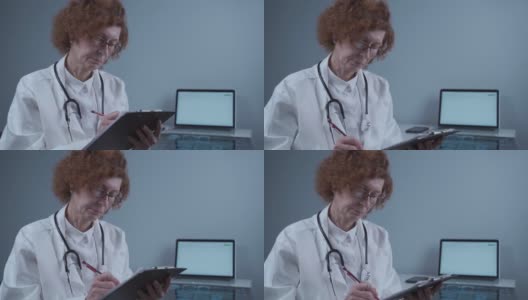 成熟的女医生在诊所办公室，在剪贴板上写医疗报告。老年女性妇科医生填写医疗文件，病历，病史，处方高清在线视频素材下载