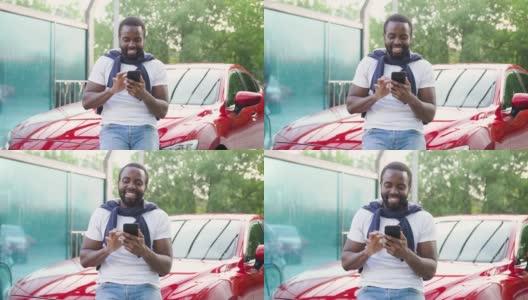 英俊微笑着留着胡子的黑皮肤年轻人站在他自己的红色名车旁洗车，享受着手机上的搞笑照片高清在线视频素材下载