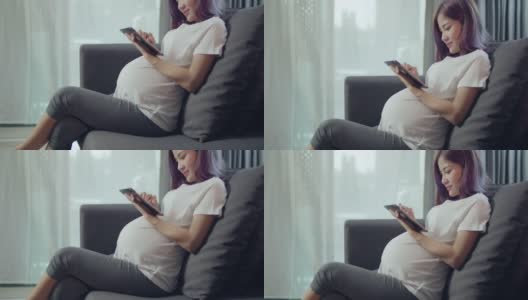美丽的亚洲孕妇抚摸肚子，而坐在沙发上的客厅在家里。高清在线视频素材下载