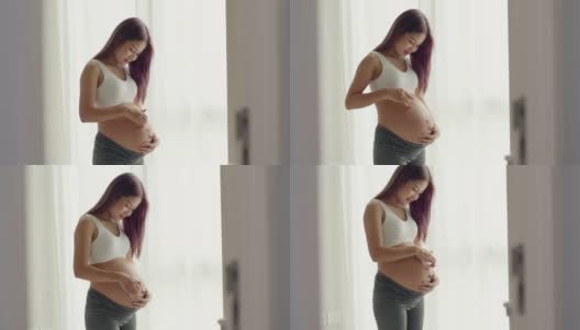 一位美丽的亚洲孕妇站在窗前，抚摸着自己的肚子。高清在线视频素材下载