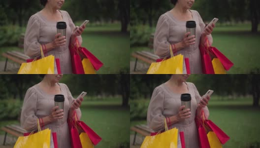 一位无法辨认的白人老年妇女拿着购物袋站在那里，用智能手机在线发短信，面带微笑。积极自信的女士拿着保温瓶在户外上网。高清在线视频素材下载