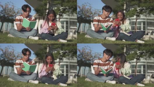 两名大学生坐在大学校园的草地上一起学习读书，用手机在社交媒体上分享快乐时光高清在线视频素材下载