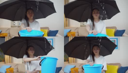 一名困惑的女子坐在客厅的伞下，拿着水桶准备接天花板漏水的水高清在线视频素材下载