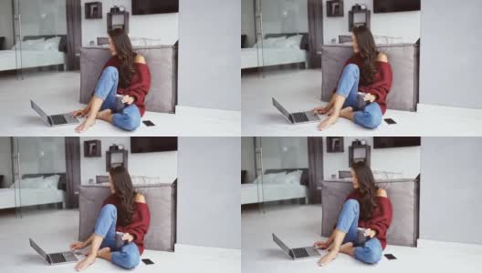微笑的黑发女人在毛衣坐在地板上的咖啡和智能手机，而使用笔记本电脑高清在线视频素材下载
