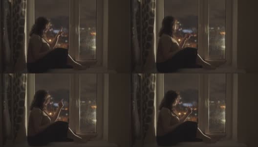 一个女人坐在窗台上，靠近潮湿的窗户。女孩用手指划过玻璃。高清在线视频素材下载