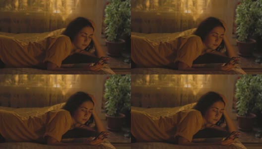 一个漂亮的年轻女人正躺在床上，靠在她的胳膊上，用着智能手机。窗外的晚霞，傍晚高清在线视频素材下载