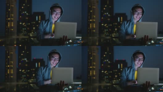 亚洲男性穿着夹克和眼镜，休闲的布料，工作类型的数据编码程序员与笔记本电脑在夜间工作，与散景灯光从城市建筑的城市景观背景高清在线视频素材下载