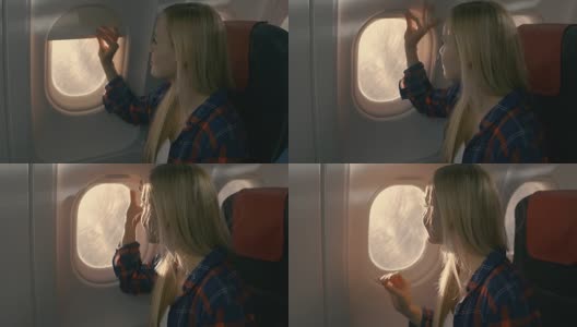 在飞机上，美丽的金发女孩打开窗帘向窗外望去。阳光照耀着她，她微笑着。高清在线视频素材下载