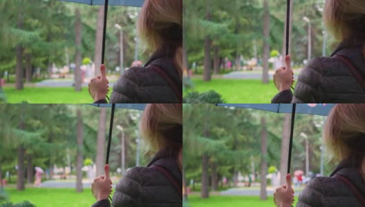 一个女人在雨天的公园里，手里拿着一把伞。一个不确定年龄的金发女人穿着一件湿夹克的后视图。在恶劣的天气里，一个不认识的女人撑着伞站在城市里高清在线视频素材下载