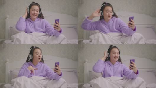 亚洲妇女在无线耳机唱歌使用手机音乐应用程序。穿着紫色睡衣的快乐女人，坐在床上手舞足蹈。高清在线视频素材下载