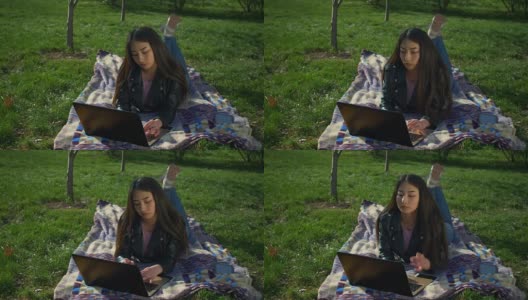 时髦女孩躺在草坪上用笔记本电脑打字高清在线视频素材下载