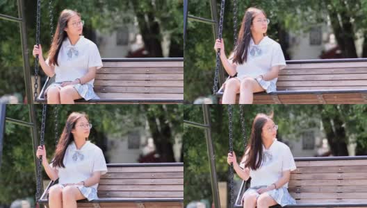 阳光明媚的日子里，可爱的中国年轻女子穿着日本学生风格的连衣裙在公园的绳梯上摇摆，戴着眼镜的漂亮女孩享受夏天的时光，超慢的动作，背光镜头。高清在线视频素材下载