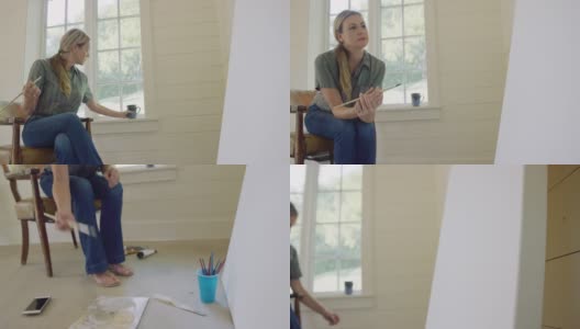 在她的室内艺术工作室里，一位四十多岁的白人女艺术家坐在椅子上，喝着咖啡，用画笔蘸着颜料，凝视着一张巨大的空白画布高清在线视频素材下载