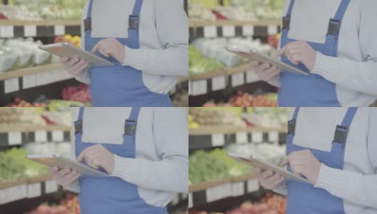 穿着制服的杂货店男员工在货架上走着，用着平板电脑。成年人在零售店检查水果和蔬菜的清单。商业、消费、生活方式。S-log 2。高清在线视频素材下载