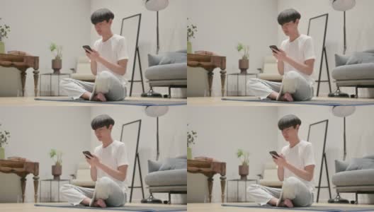 年轻的亚洲男子在瑜伽垫上使用智能手机高清在线视频素材下载