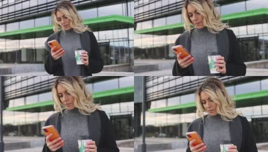 年轻漂亮的女商人喝着咖啡在办公大楼前用手机发短信高清在线视频素材下载