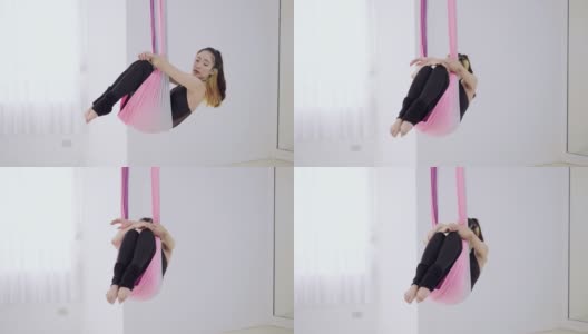 运动的亚洲妇女在吊床上做瑜伽姿势的特写肖像。用反重力材料吊在绳子上的女人。迷人的运动女人做空中瑜伽练习，放松地坐在彩色吊床上。空中体操，杂技和反重力高清在线视频素材下载