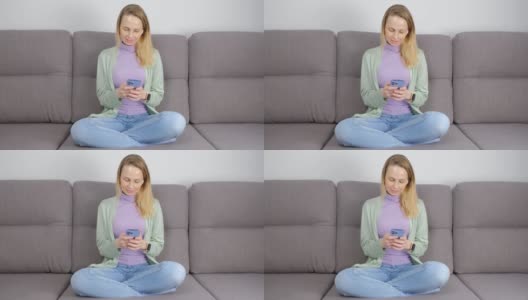 一名悠闲的白人女子，盘腿坐在家里的沙发上，用智能手机打字，戴着无线耳机听音乐。美丽的女性用手机在线交流高清在线视频素材下载