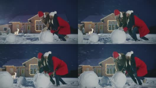 快乐的年轻夫妇堆雪人，都滚大雪球。一家人在一个冬天的晚上玩耍。在装饰着花环的背景房子里。高清在线视频素材下载