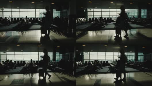 一位女士提着带轮子的包，手里拿着手机正在机场候机楼里走着。大窗户映衬下的剪影高清在线视频素材下载