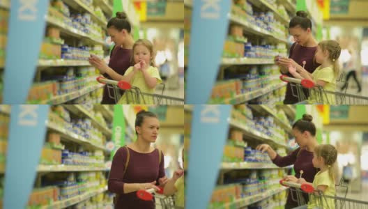 可爱的女孩与白人外表是吃棒棒糖和坐在超市手推车。背景母亲为女儿选择婴儿食品高清在线视频素材下载