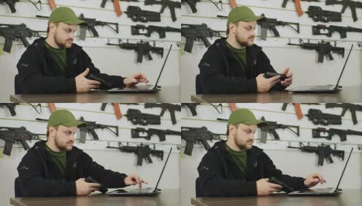 一个枪支贩子在工作。坐在商店的电脑前，在架子的背景上有不同的武器高清在线视频素材下载