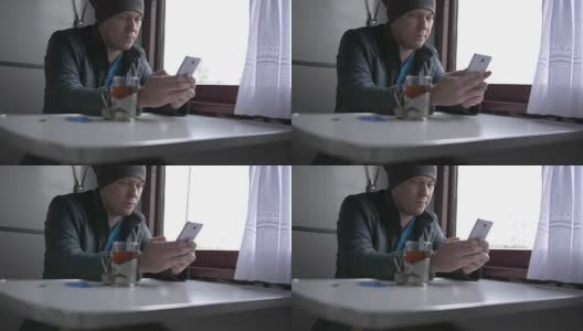 一位戴着灰色帽子、穿着蓝色夹克的年轻人手持白色智能手机与人交流，微笑着将智能手机放在桌子上，然后端上一杯茶高清在线视频素材下载