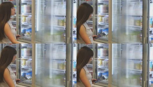 年轻女子在超市的冷藏区打开玻璃门购买乳制品或冷藏食品。女孩在商店从冰箱里拿东西，用平板电脑检查购物清单高清在线视频素材下载