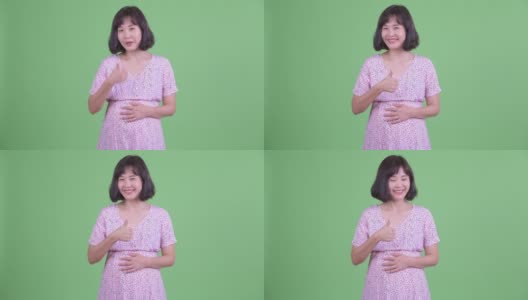 摄影棚拍摄了一名美丽的亚洲孕妇，在绿色的摄影棚背景下微笑并竖起大拇指高清在线视频素材下载