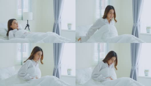 亚洲美丽生病的女孩在睡衣起床在卧室。有魅力的年轻女性在床上醒来后会感觉很难受，肚子疼或者早上抱肚子。高清在线视频素材下载