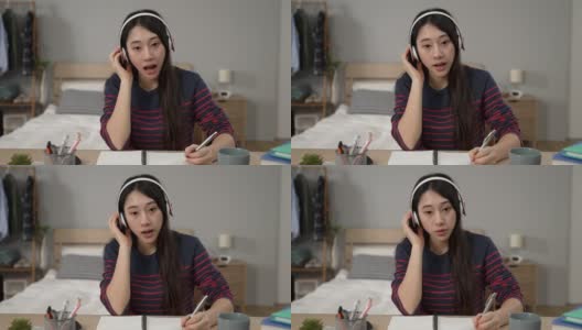 戴着耳机在家学习的亚洲女性学生的正面形象是在网上上课时重复句子和写笔记高清在线视频素材下载