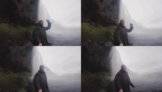 一个快乐的年轻人拿着自拍杆在大雾天用gopro相机拍摄强大的Seljalandsfoss瀑布高清在线视频素材下载