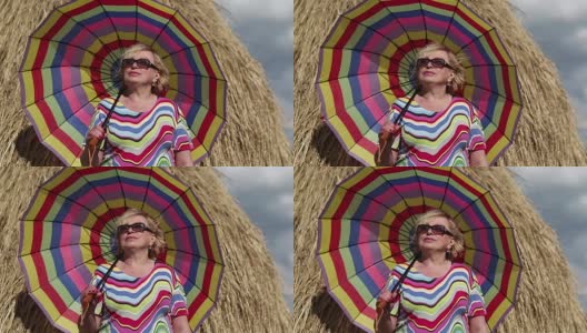 一个戴着眼镜，打着彩色雨伞的金发女人呆在干草堆附近高清在线视频素材下载