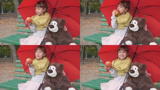 孩子坐在木凳上，旁边是一把红色伞下的泰迪熊，在公园里吃大红高清在线视频素材下载