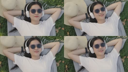 美丽的年轻亚洲女人戴着太阳镜和耳机睡在草地上。高清在线视频素材下载