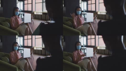 亚洲妇女戴着口罩，坐在沙发上使用笔记本电脑在现代办公室高清在线视频素材下载