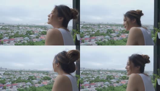 年轻美丽的亚洲女人在早上站在公寓阳台上，欣赏大都市的城市景观建筑的景色。2019冠状病毒病大流行封锁和心理健康概念。高清在线视频素材下载