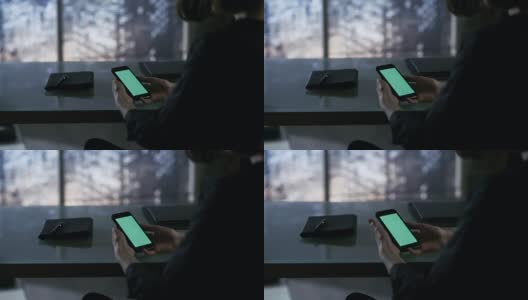 肩上的女商人坐在她的办公桌上，使用绿色屏幕的智能手机。从窗外看大城市。高清在线视频素材下载