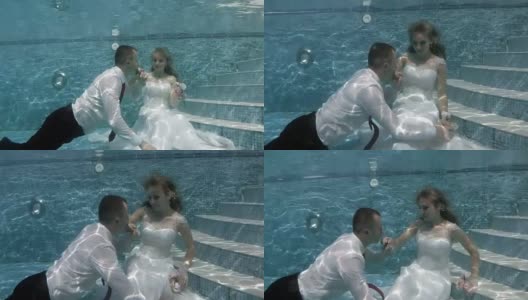 幸福的新郎和新娘穿着白色婚纱坐在池底。新郎飘起来，亲吻新娘的手高清在线视频素材下载