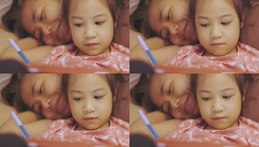 亚洲妈妈和可爱的女儿躺在家里的地板上，在周末用科技娱乐的平板电脑上书写快乐快乐的时刻。高清在线视频素材下载