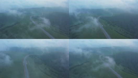 从山顶上可以看到穿过山丘的山路。无人机在绿色田野上空的云层中飞行高清在线视频素材下载