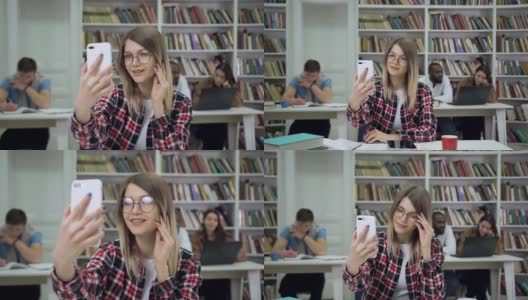 美丽微笑的现代明亮的女学生戴着眼镜坐在图书馆里，看着手机摄像头上其他人多样化的学生背景高清在线视频素材下载