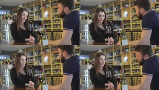 一个男人和一个女人正在用玻璃杯喝红酒，一个餐厅的内部，一个年轻的男人和一个女孩在一个餐厅的桌子上喝葡萄酒，背景是一个葡萄酒架子，一个男性嬉皮士高清在线视频素材下载