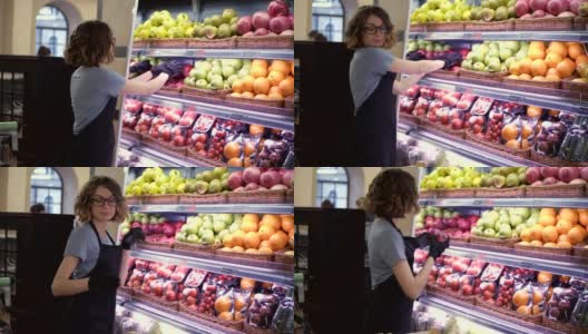 近距离观察漂亮的白人工人在黑色围裙和手套在超市储存水果。戴眼镜的年轻员工。卷曲的女性在架子上排列着红色的大苹果。慢动作高清在线视频素材下载