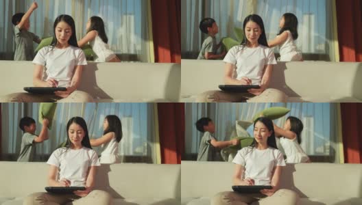 亚洲母亲喜欢把注意力集中在平板电脑上，在家工作。她的儿子和女儿在后面玩枕头大战。为了欢乐的家庭时光高清在线视频素材下载