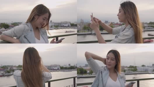 年轻美丽的亚洲女游客站在阳台上，用智能手机相机自拍或在日落时拍摄美丽的城市和河流。美丽的女孩放松，享受户外生活方式，度假旅行的城市。高清在线视频素材下载