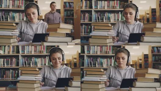 一位戴着白色耳机、面带微笑的年轻女子正坐在大学图书馆的桌子旁，手里拿着平板电脑，听着音乐。她在跳舞，在笔记本上写讲座高清在线视频素材下载