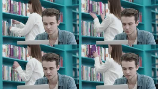 男生在大学图书馆专注地用笔记本电脑做作业，女生在后台浏览书架上的书高清在线视频素材下载